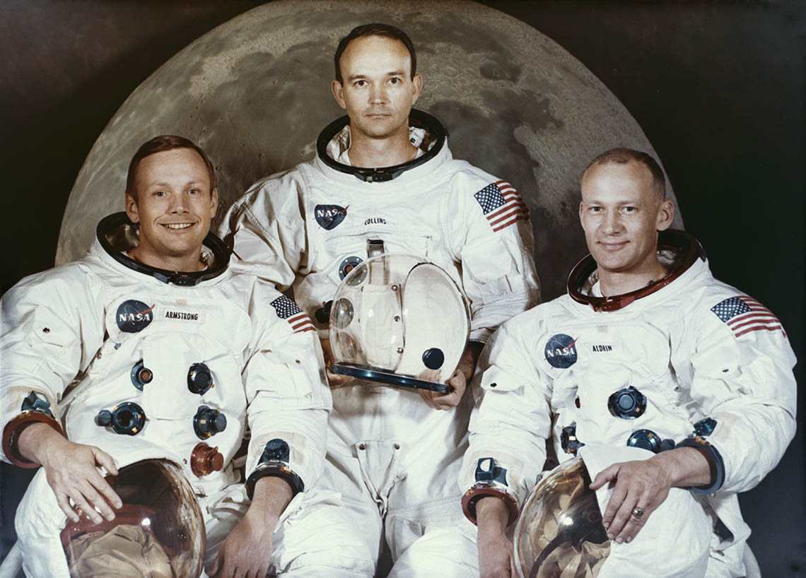 Three white male astronauts in flight suits posing in front of a picture of the moon.