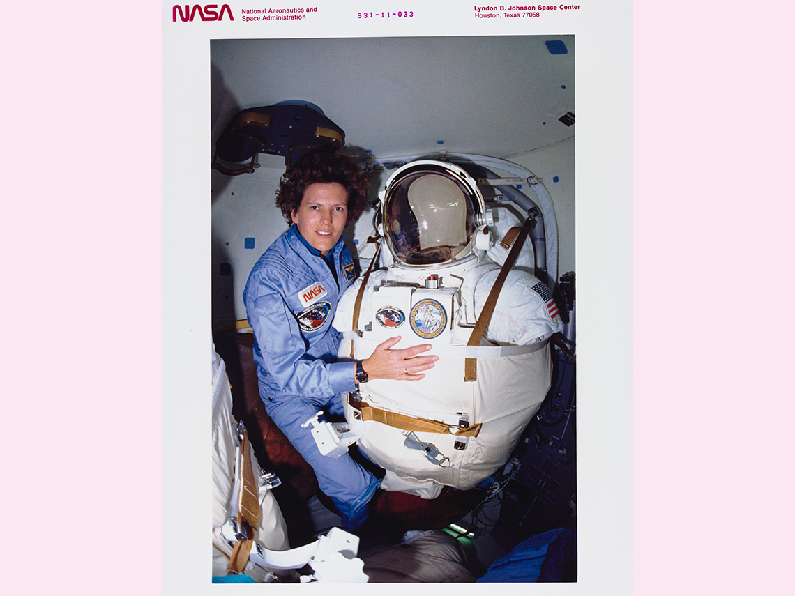 A white female NASA astronaut in an airlock, ready to put on the suit that will let her walk in space.