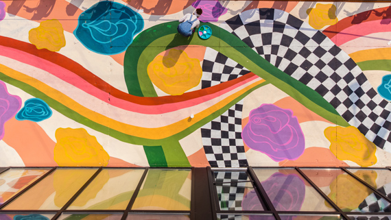 A giant mural of colourful flowers and winding ribbons being painted by a person onto a sidewalk