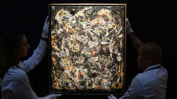 An abstract painting made of thick textured layers of chaotic drips and streaks being held by art technicians in protective gloves