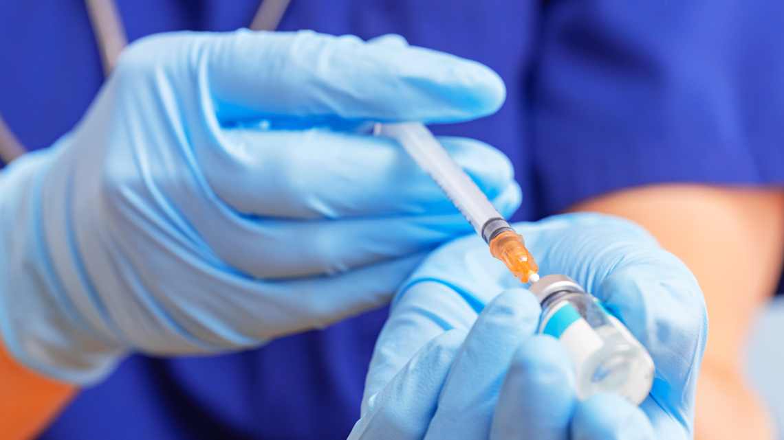 A person wearing medical scrubs and protective gloves filling a syringe with a substance from a vile