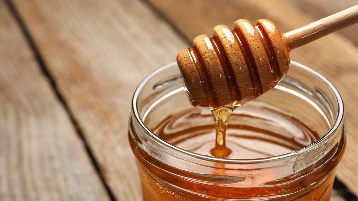 A jar of fresh honey, with a wooden honey dipper shaped like a honeycomb in the jar with honey dripping off of it.