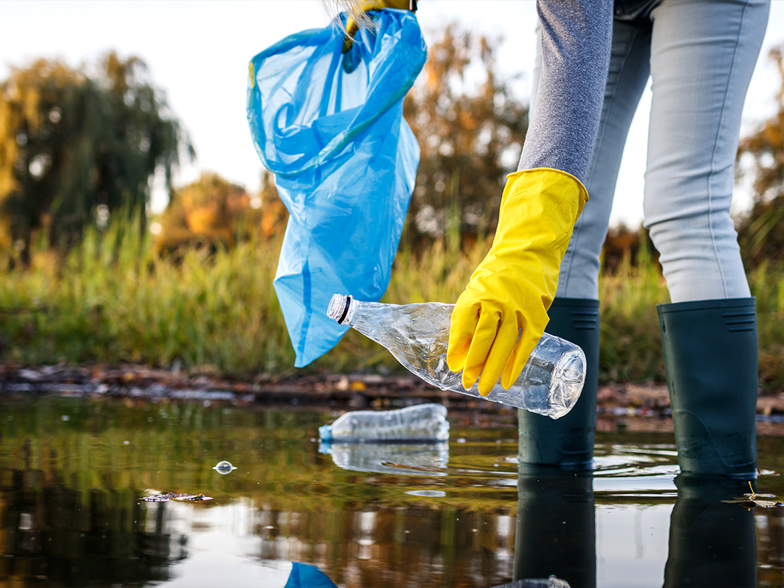 A person wearing yellow rubber gloves picks up a plastic bottle from a lake.