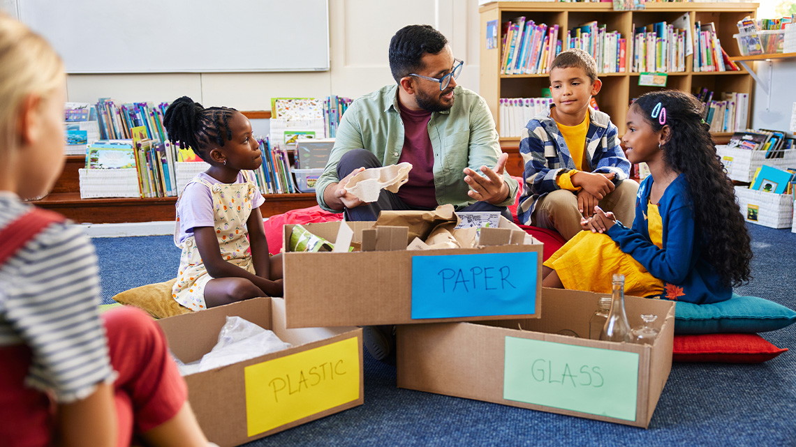 A teacher and students sitting together and sorting recyclables into boxes labeled 'paper,' 'plastic,' and 'glass.'