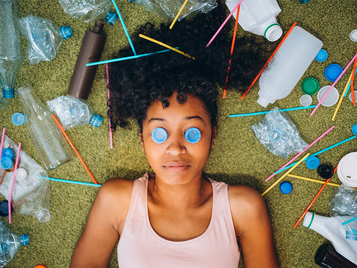 A student laying on their back on grass. They have an assortment of plastics around them and two bottle caps placed on their eyes.