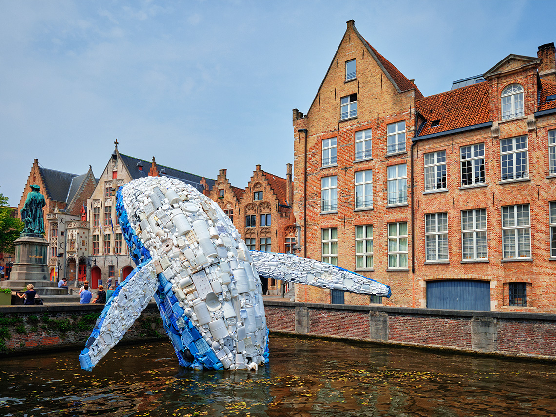 A large art piece of a whale made from 5 tons of plastic collected from the ocean. There are medium sized buildings in the background.