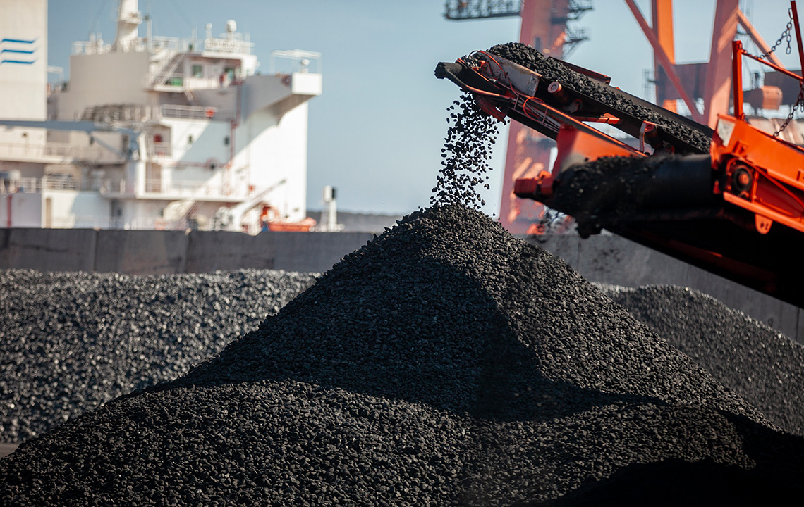 A machine dumping coal onto a large pile of coal at a port. There is a factory in the background.