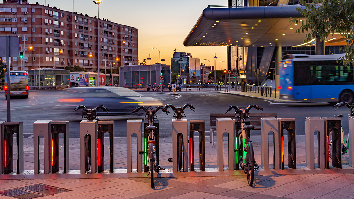 A line up of a bike rental at a bus station. There is a building and vehicles in the background.
