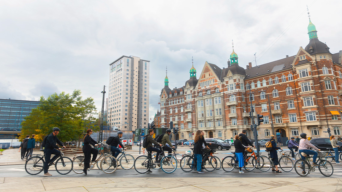 There is a crowd of people lined up with bicycles. There are three large buildings in the background.