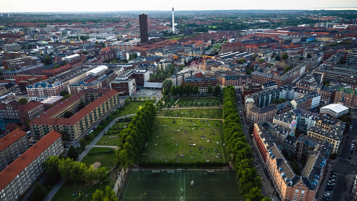 Birds eye view of multiple buildings. There is a large sports court and open land of grass surround by trees in the middle.