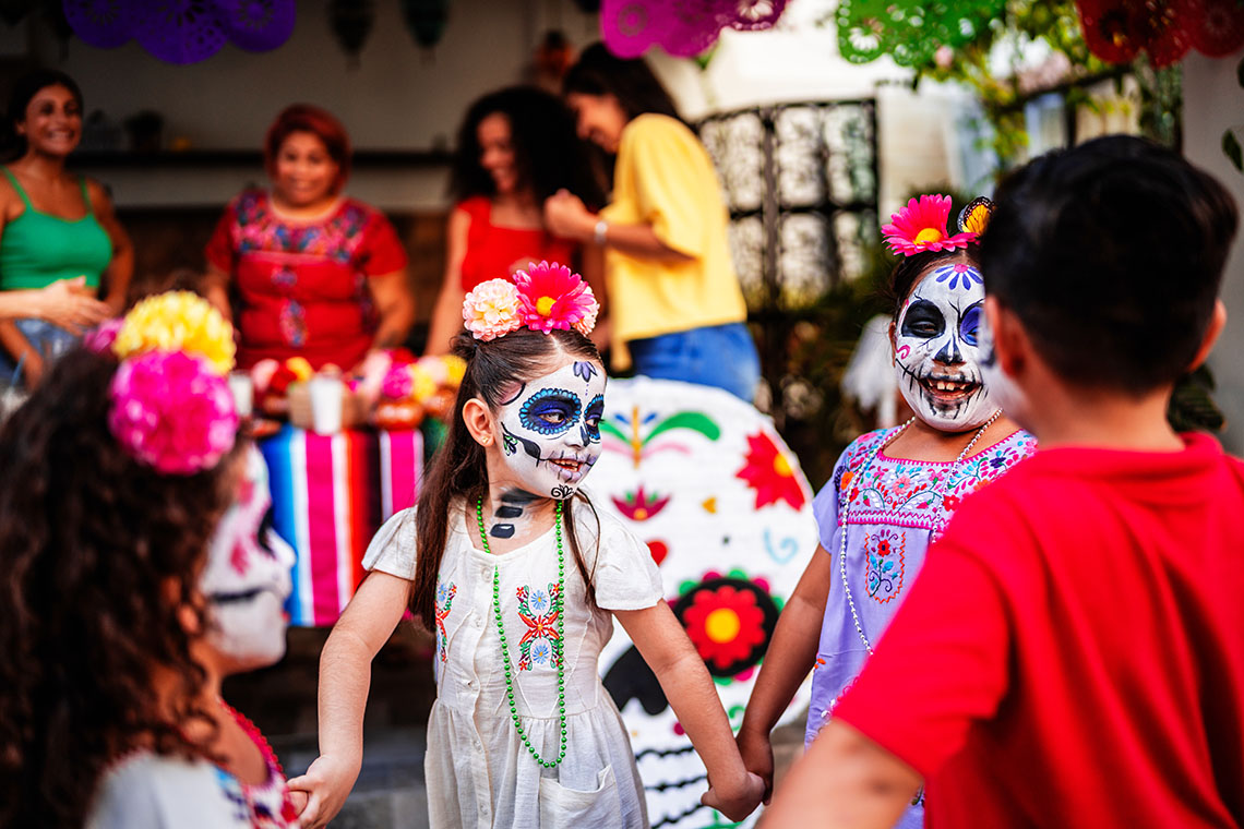 Children with faces painted holding hands during an outdoor day of the dead celebration.