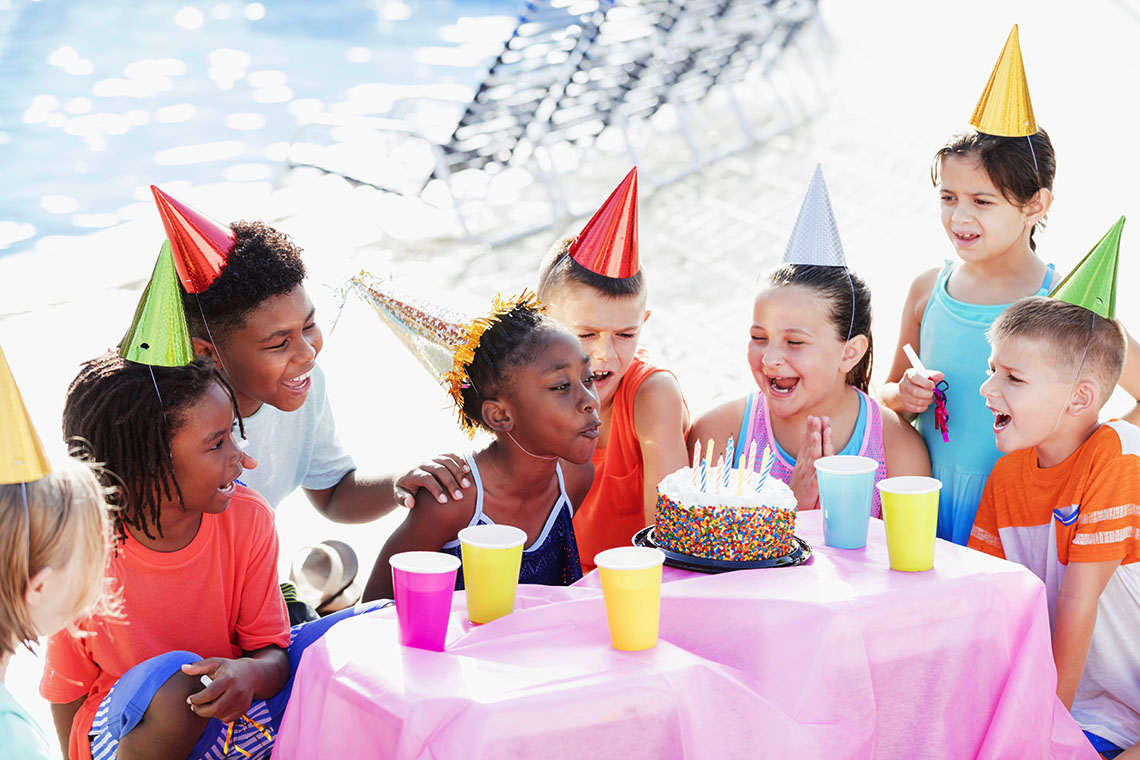 Group of children sitting outside surrounding a child who is blowing out candles from a cake.