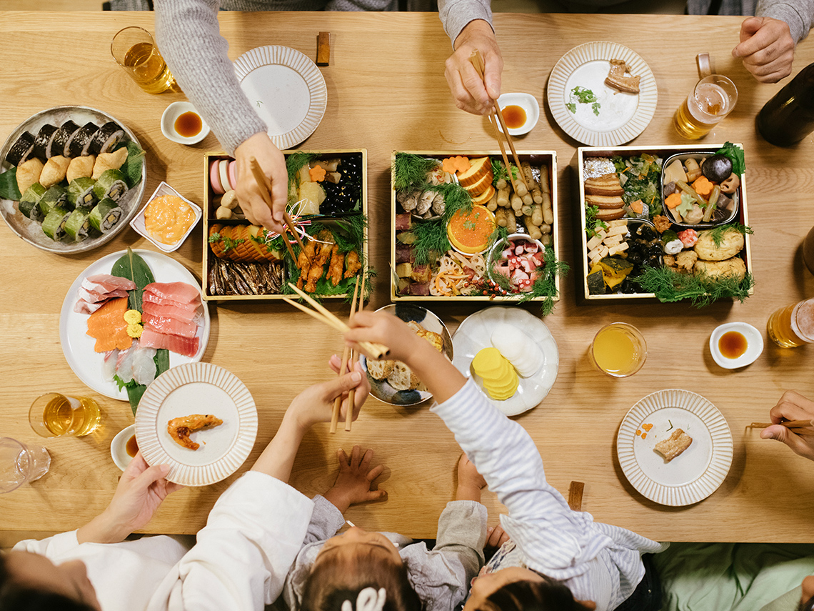A family sharing three large boxes with many different kinds of food, along with several plates of extra kinds of food.
