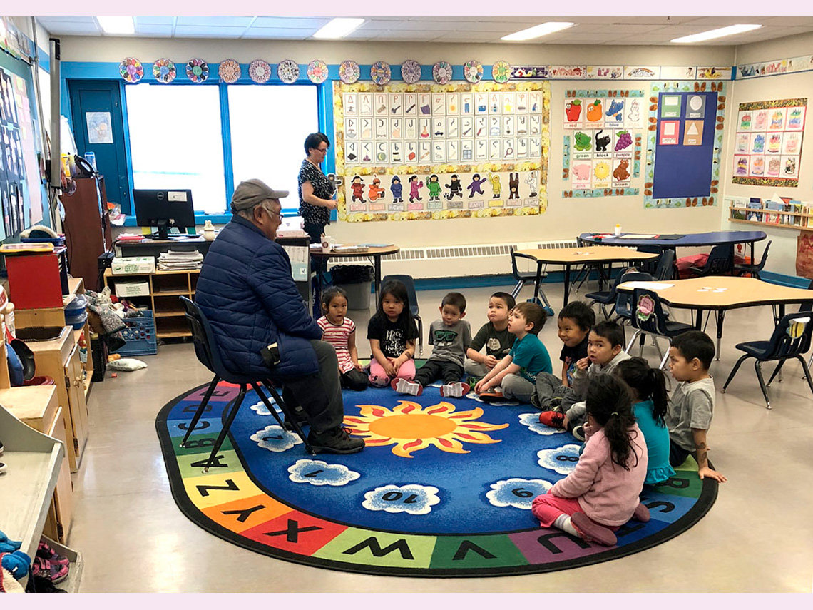 Students in their classroom sitting in a circle on the carpet facing an Elder seated in a chair.
