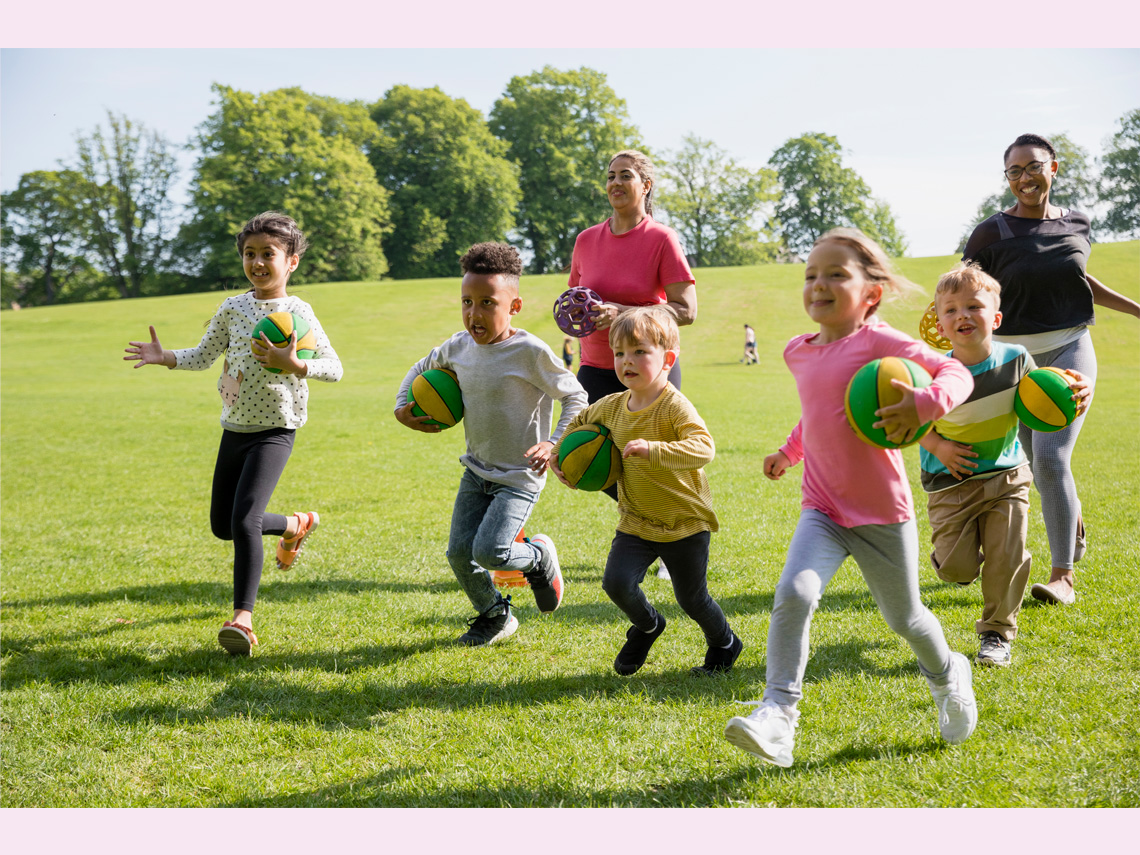 A team of children run across a field holding balls. Two adults are behind them with different kinds of sports balls.