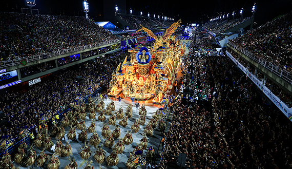 A huge stadium of people are watching a Carnival parade with many dancers performing and very large decorated floats moving down the stage.