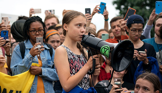 Greta Thunberg speaking into a megaphone surrounded by youth recording with their phones