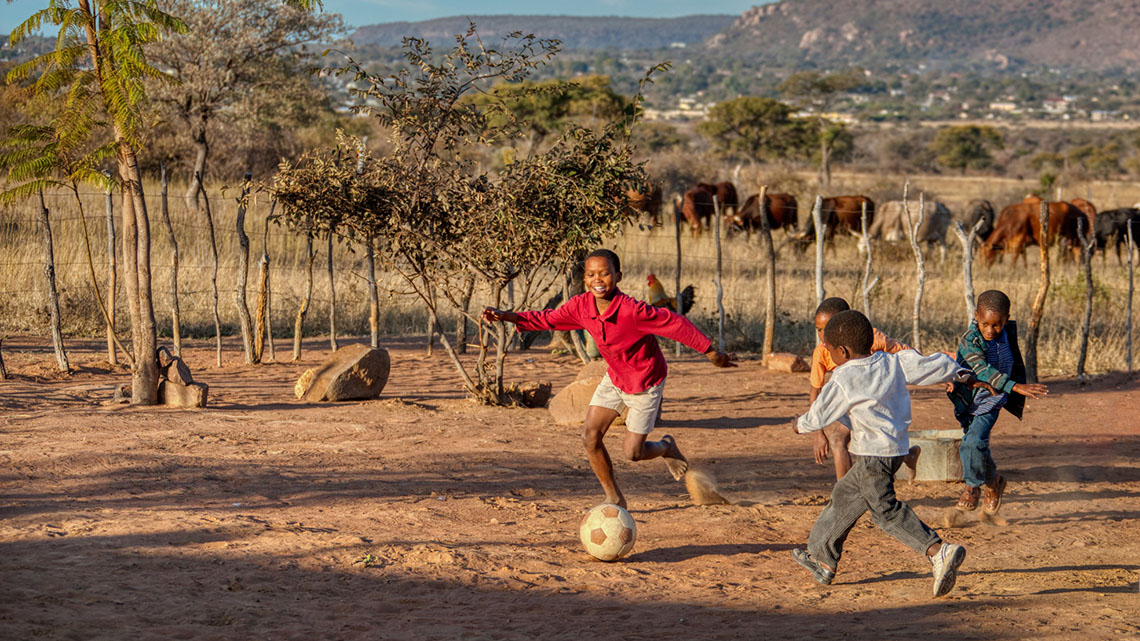 Smiling children wearing long-sleeve shirts, pants, and shorts kicking a ball in a dirt yard with savannah plants and trees