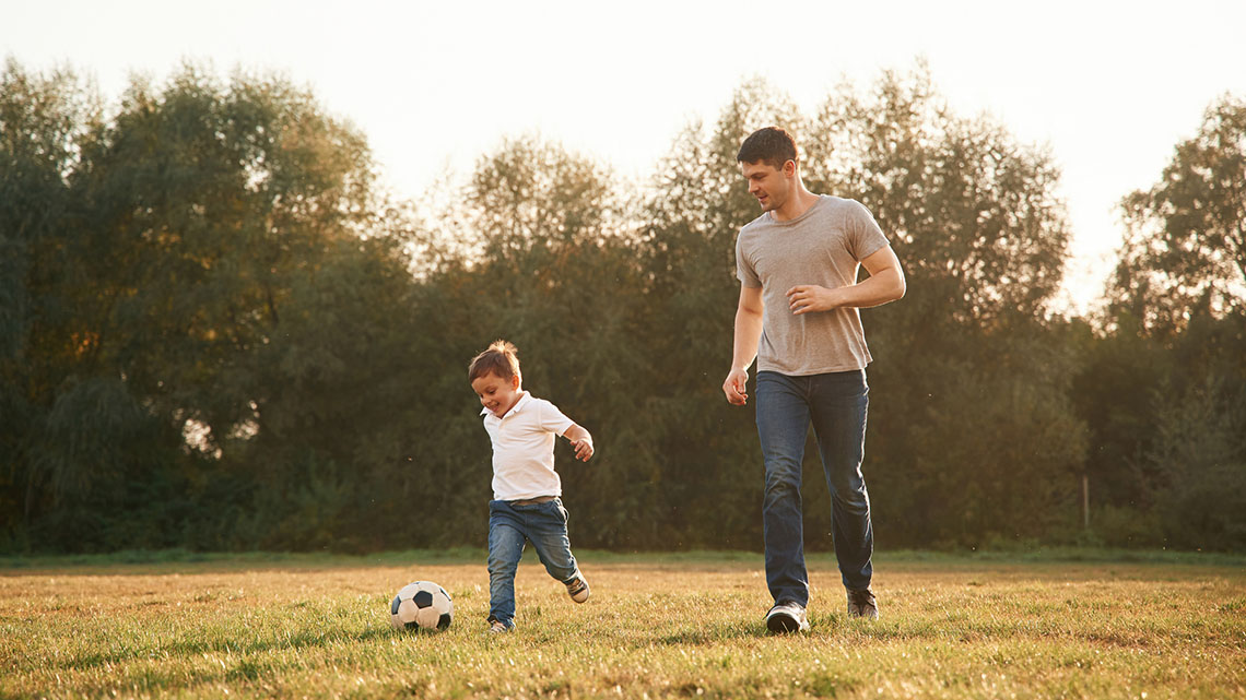 A smiling child wearing a short-sleeve shirt and pants kicking a ball in a grass field with temperate plants and trees