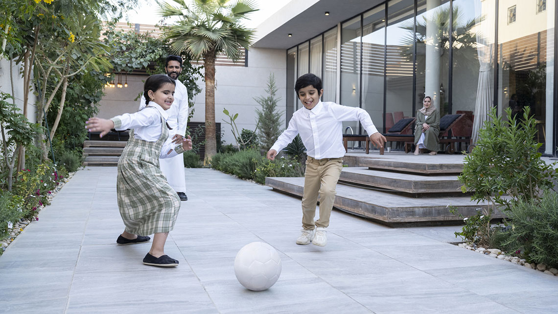 Smiling children wearing long-sleeve shirts, pants, and long skirts kicking a ball in a courtyard with mediterranean plants and trees