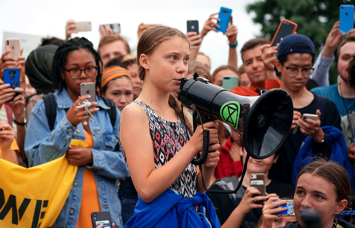 Greta Thunberg speaking into a megaphone surrounded by youth recording with their phones