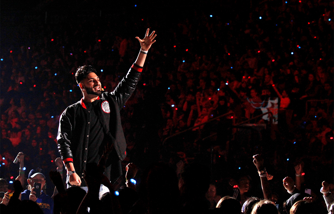 Wali Shah raising his hand and speaking into a headset microphone in an arena full of people