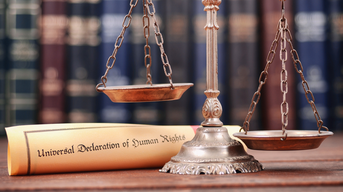 A piece of paper reads, ‘Universal Declaration of Human Rights.’ It is next to a set of scales which are unbalanced and not equal.