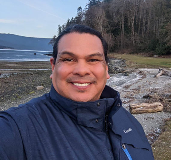 Rueben George standing on a shoreline next to a forest