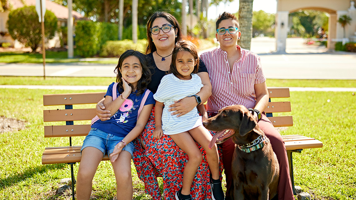 A family with two parents, two children, and a dog sitting together on a park bench and smiling.