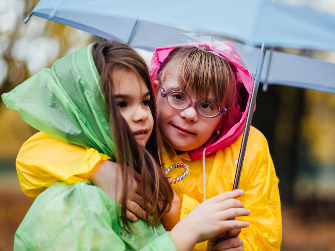 Two children wearing raincoats and sharing an umbrella in the rain.