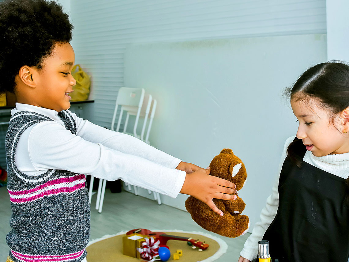A child extending their arms to share a toy with another child while playing.