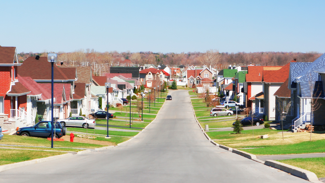 A road going through a residential neighbourhood. Each house has a grassy lawn and a driveway with a car parked in it.