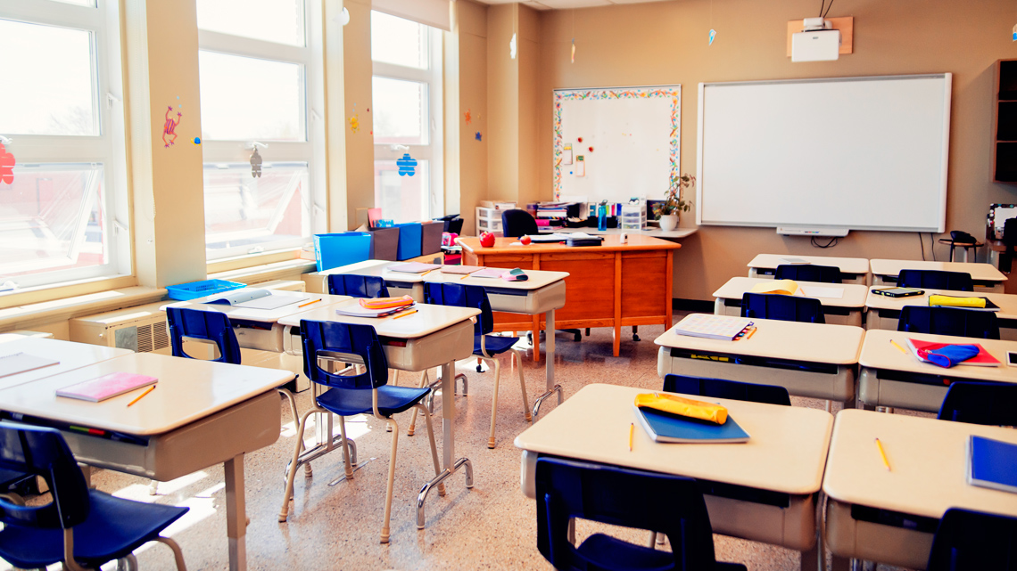 An empty classroom with rows of desks and chairs, notebooks on the desks, a whiteboard at the front, and a teacher's desk.