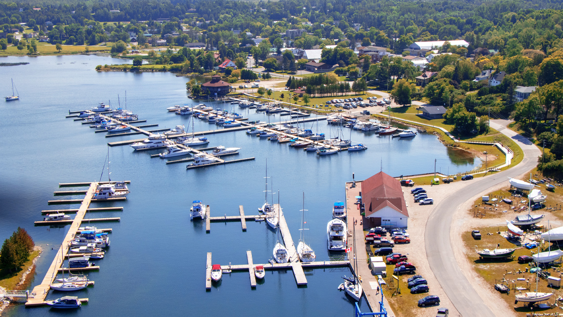 A scenic town with lush trees and large homes located next to a bay. There are many boats in the marina and parked on land near shore.