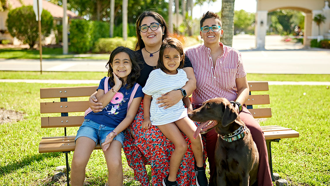 A family with two parents, two children, and a dog sitting together on a park bench and smiling.