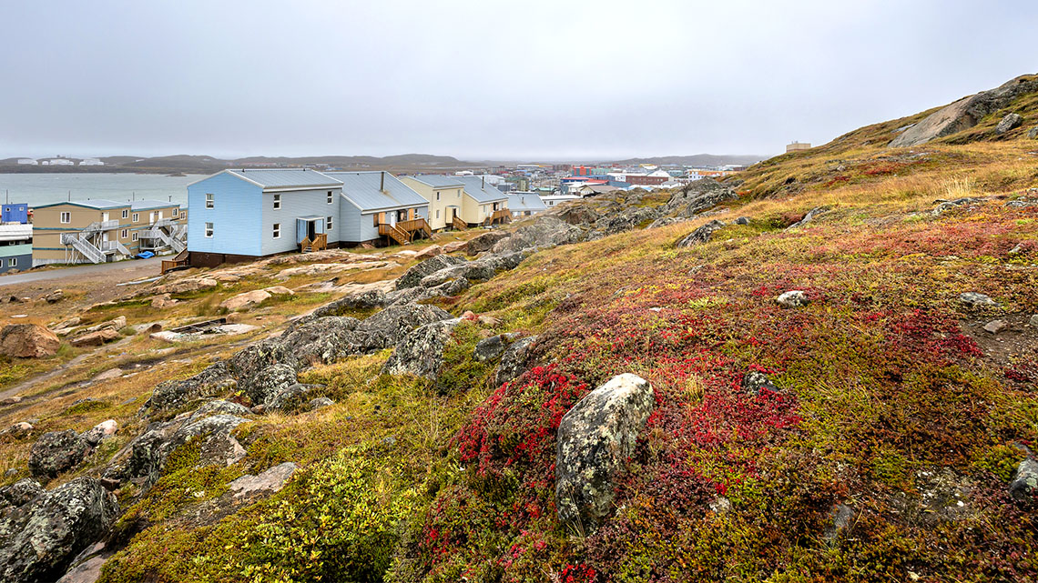 A small community of one and two story buildings next to a lake. The surrounding landscape is covered in low plants, with no trees around.
