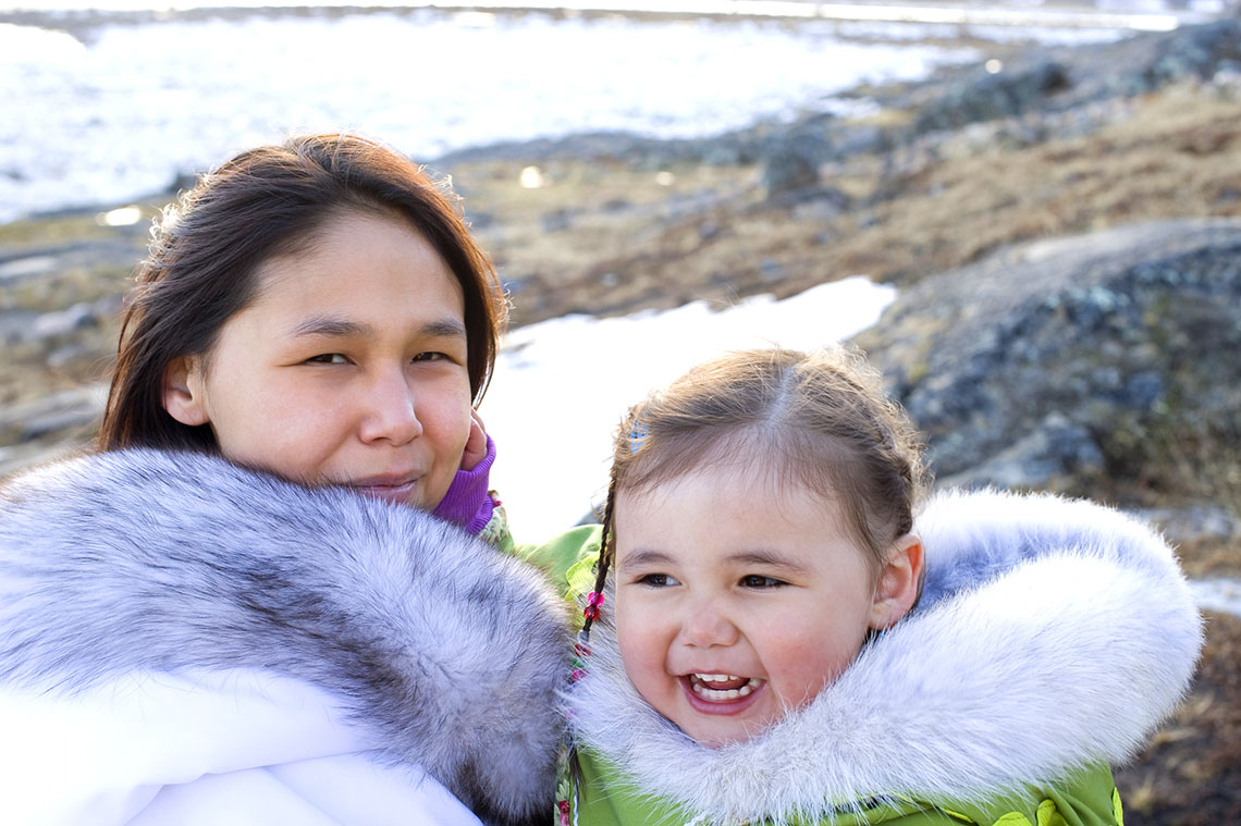 A mother and daughter are outside. They are both wearing winter jackets with hoods trimmed in thick fur.