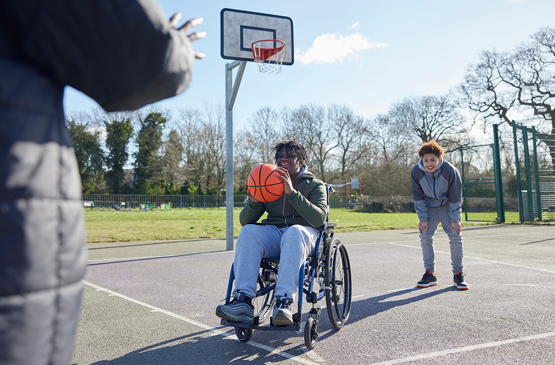 A group of kids playing basketball together at recess. One child uses a wheelchair and is throwing the ball towards the basket.