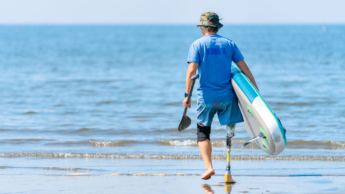 A man with a prosthetic leg, holding a surfboard while walking towards the ocean.