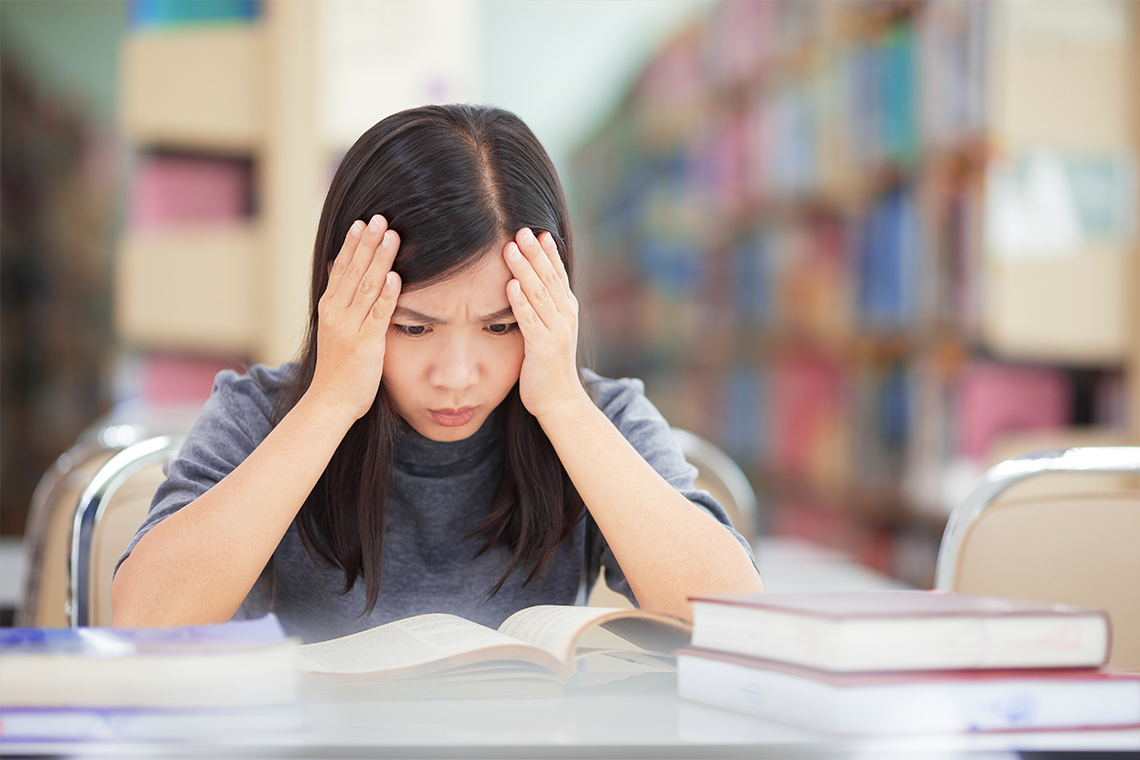 A student looking at a book while holding her head with her hands. A pile of books is visible on the desk.