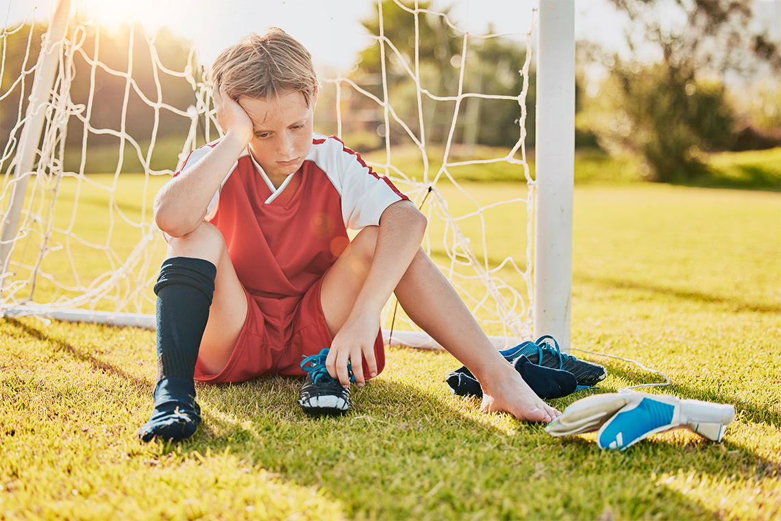 A student sits on a soccer ball in the corner of a football field, gazing down at the ground with a troubled expression. The student is wearing a soccer uniform.