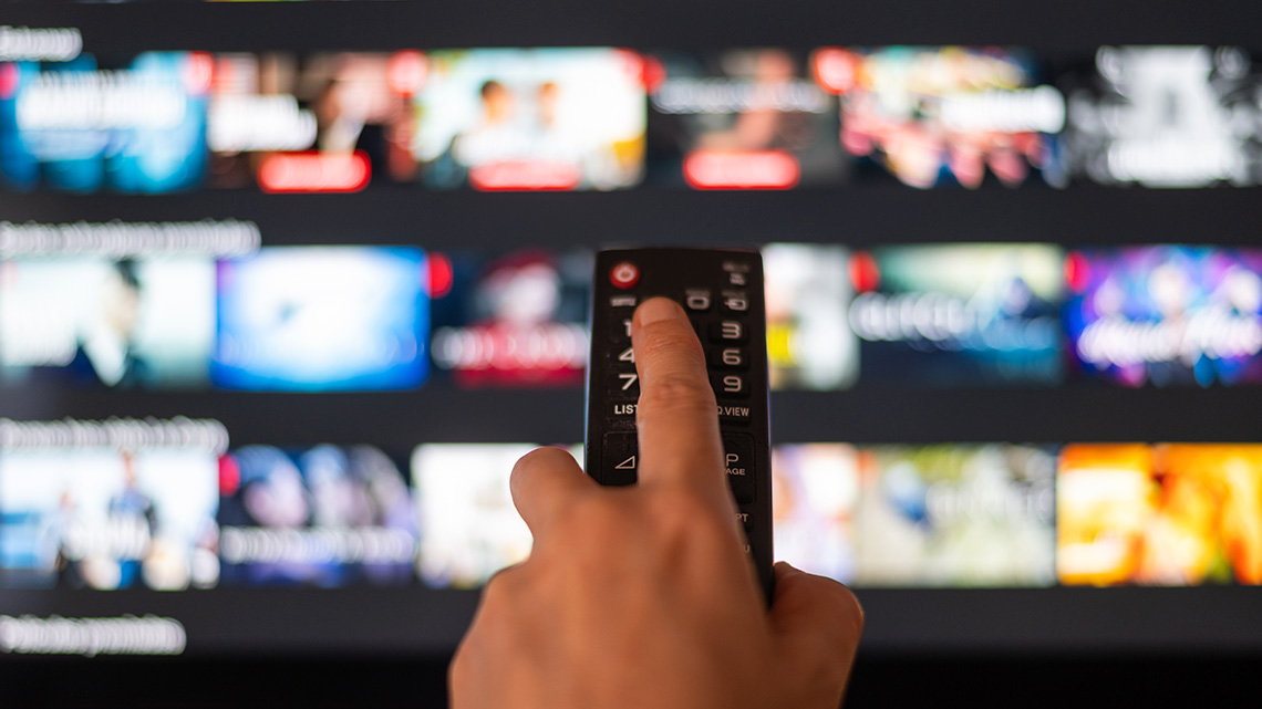 A hand holding a remote control pointed at a large screen. The screen shows various tv and movie options for selection.