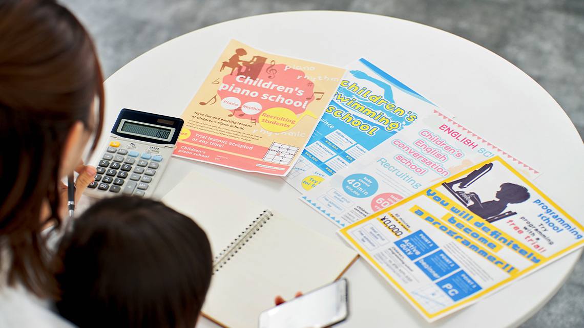 A parent sitting with a child looking at flyers for extracurricular activities. They are using a calculator and writing in a notebook