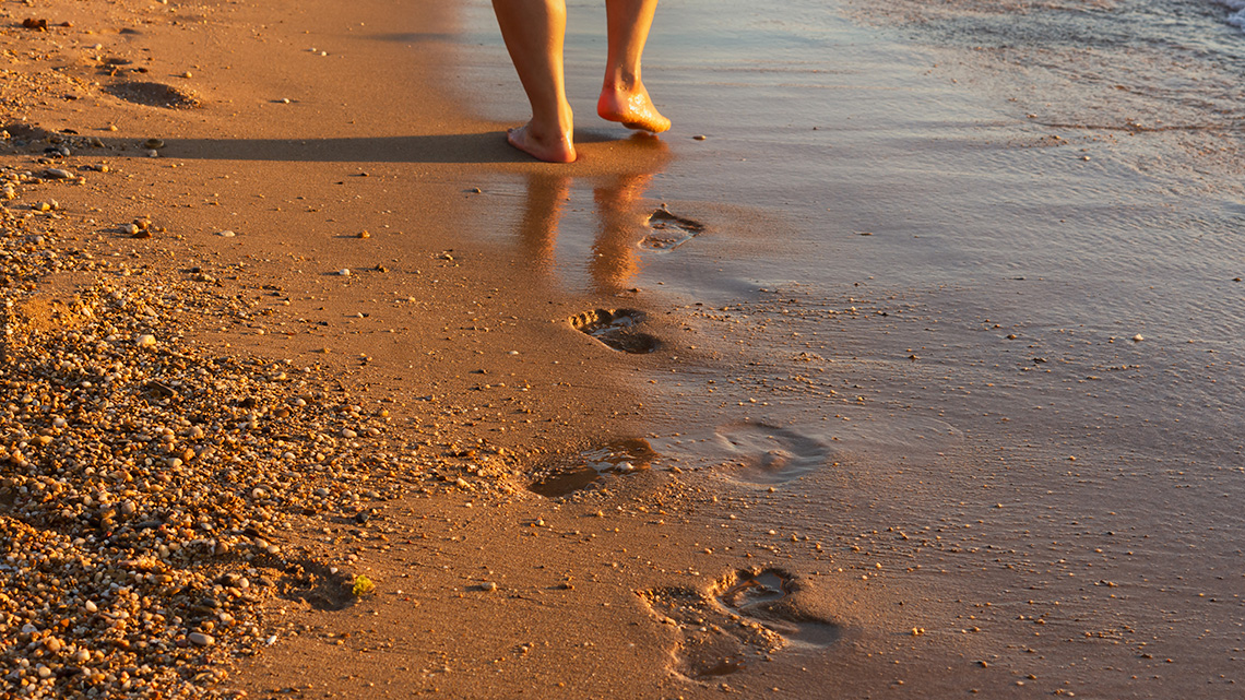 A person walking on a wet, sandy shore, leaving a trail of footprints in the sand.