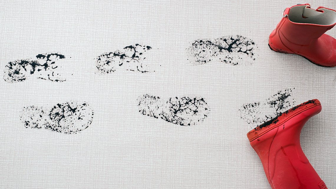 A trail of muddy footprints on a carpet leading to a pair of red rubber boots.