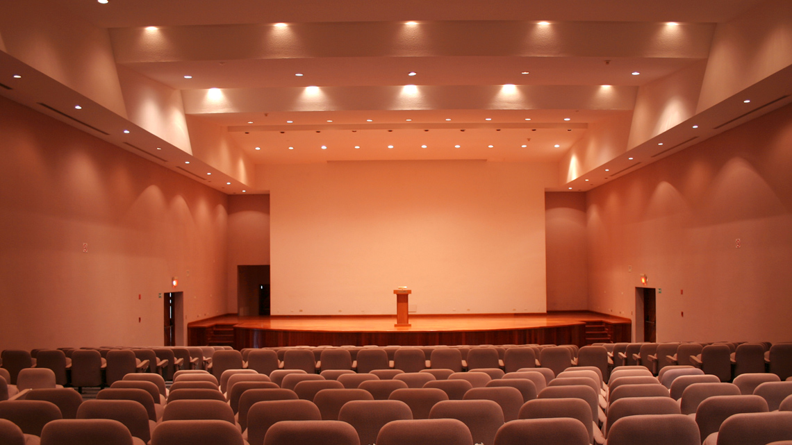 An empty auditorium with many rows of seats and a stage with a podium and a screen behind it