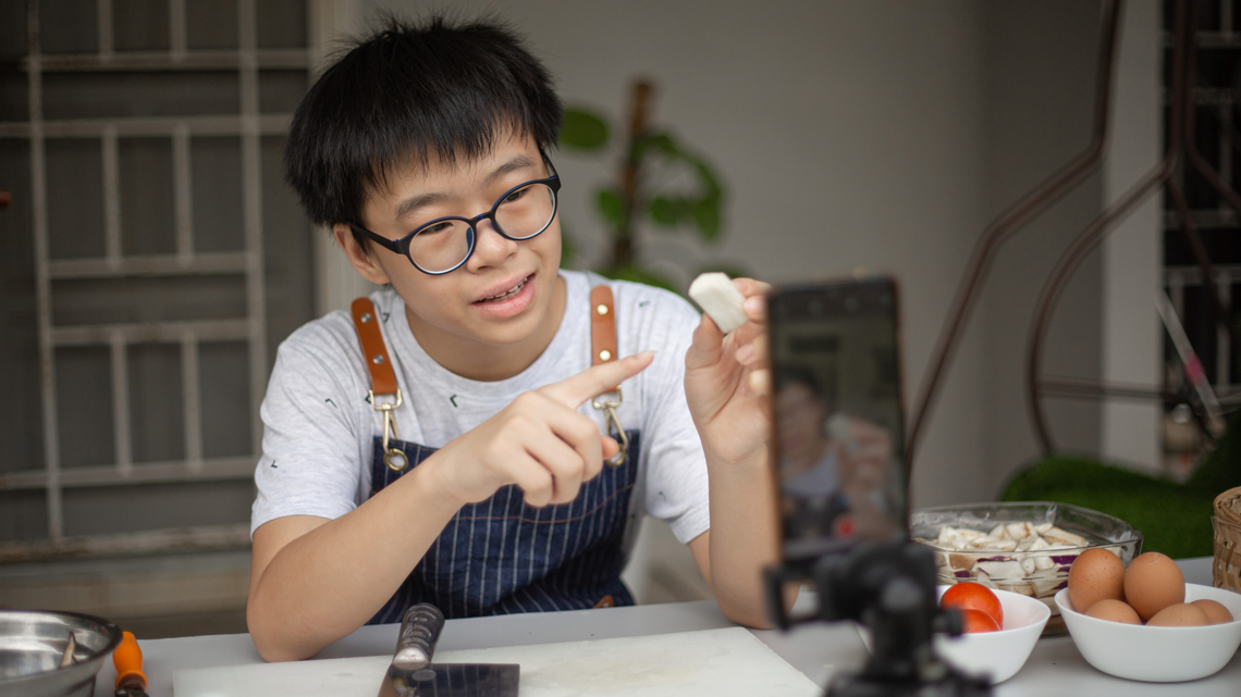 A child in a kitchen recording a video of themselves preparing food on a cellphone