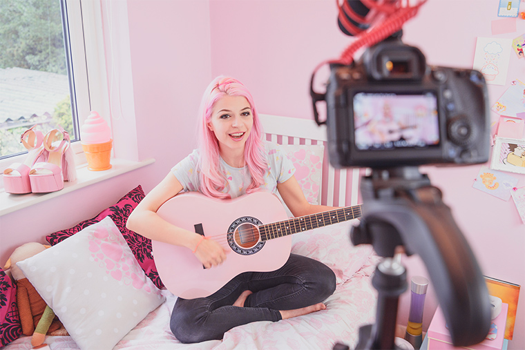 A musician sitting on a bed in a bedroom. She is playing an acoustic guitar and singing. She is filming herself with a camera on a tripod.