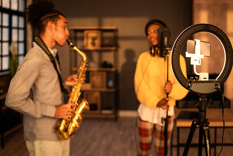 Two musicians filming themselves. There is both natural light coming in from windows, and a ring light around their phone on a tripod.