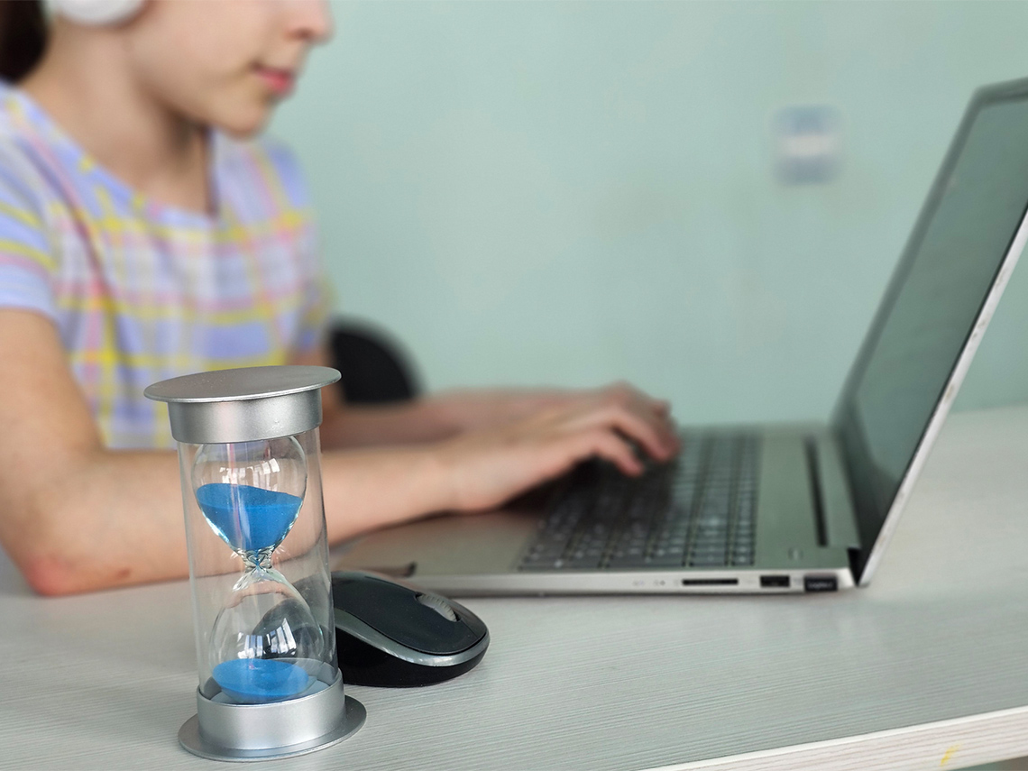 A student working on a laptop with an hourglass in the foreground.